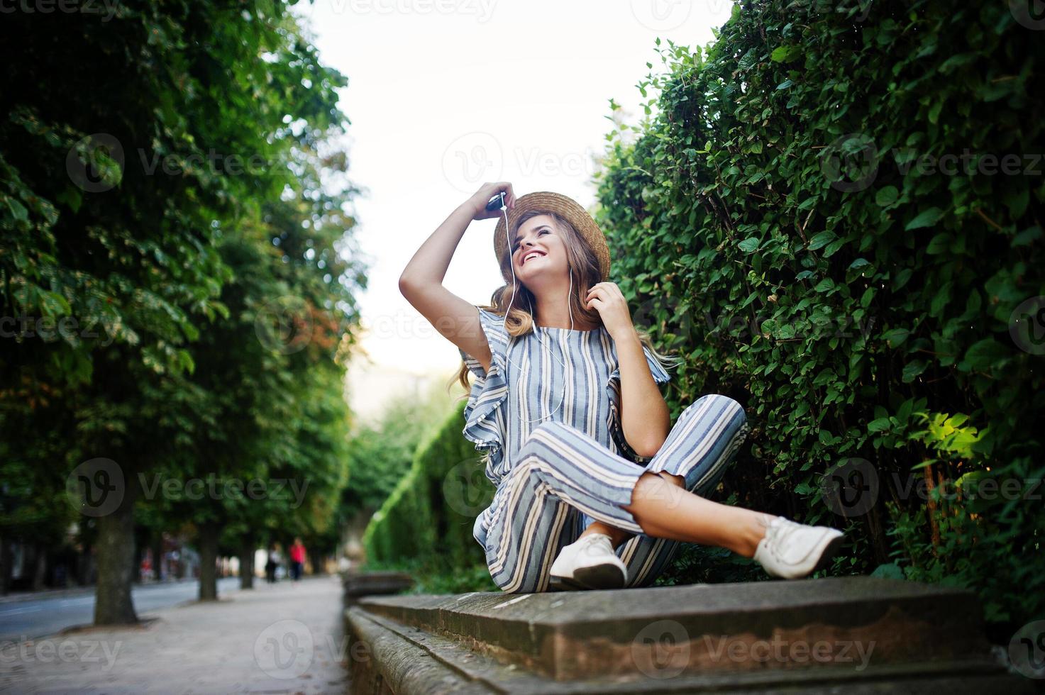 Portrait of a stunning young woman in striped overall sitting in the park and listening to the music with her earphones on. photo