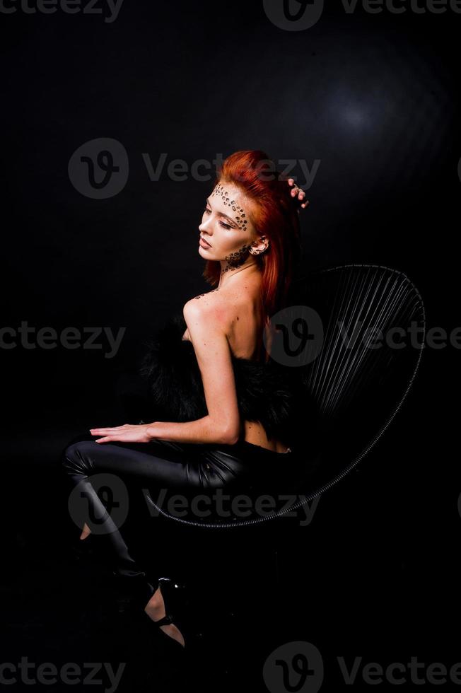 Fashion model red haired girl with originally make up like leopard predator isolated on black. Studio portrait on chair. photo