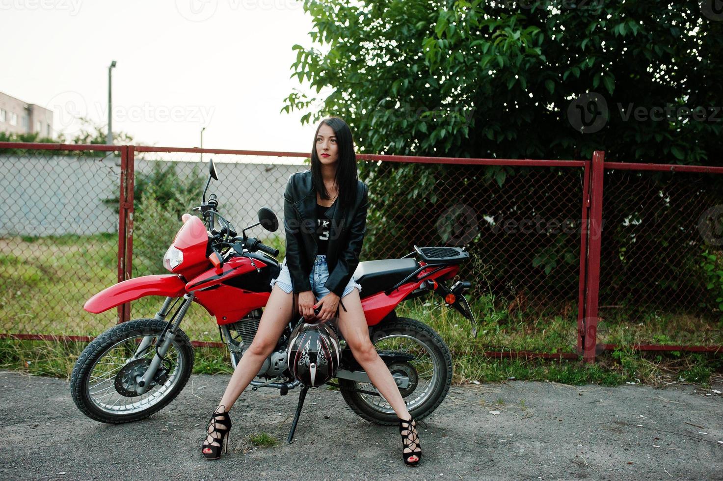 retrato de una mujer genial e impresionante con vestido y chaqueta de cuero negro sentada en una moto roja genial. foto