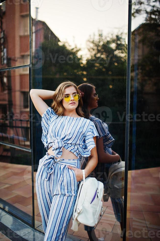 retrato de una hermosa joven con gafas de sol amarillas y a rayas posando con una mochila de cuero blanco en un balcón de un moderno edificio de vidrio en la ciudad. foto