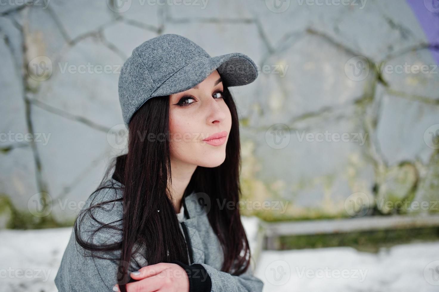 Stylish brunette girl in gray cap, casual street style on winter day against colored wall. photo