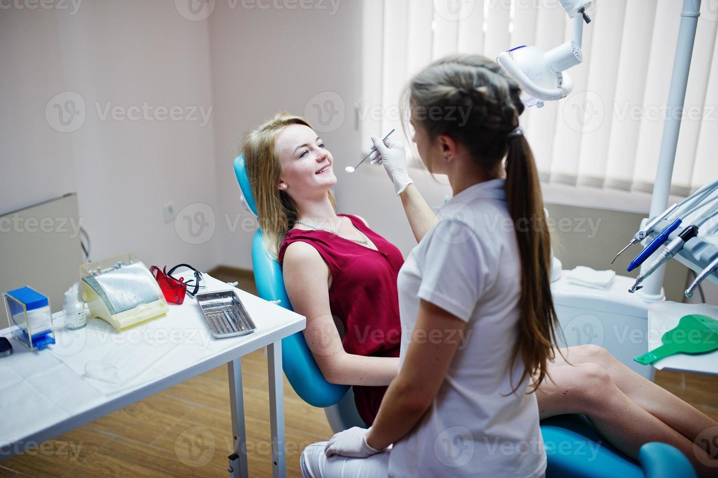 Attractive patient in red-violet dress laying on the dental chair while female dentist treating her teeth with special instruments. photo