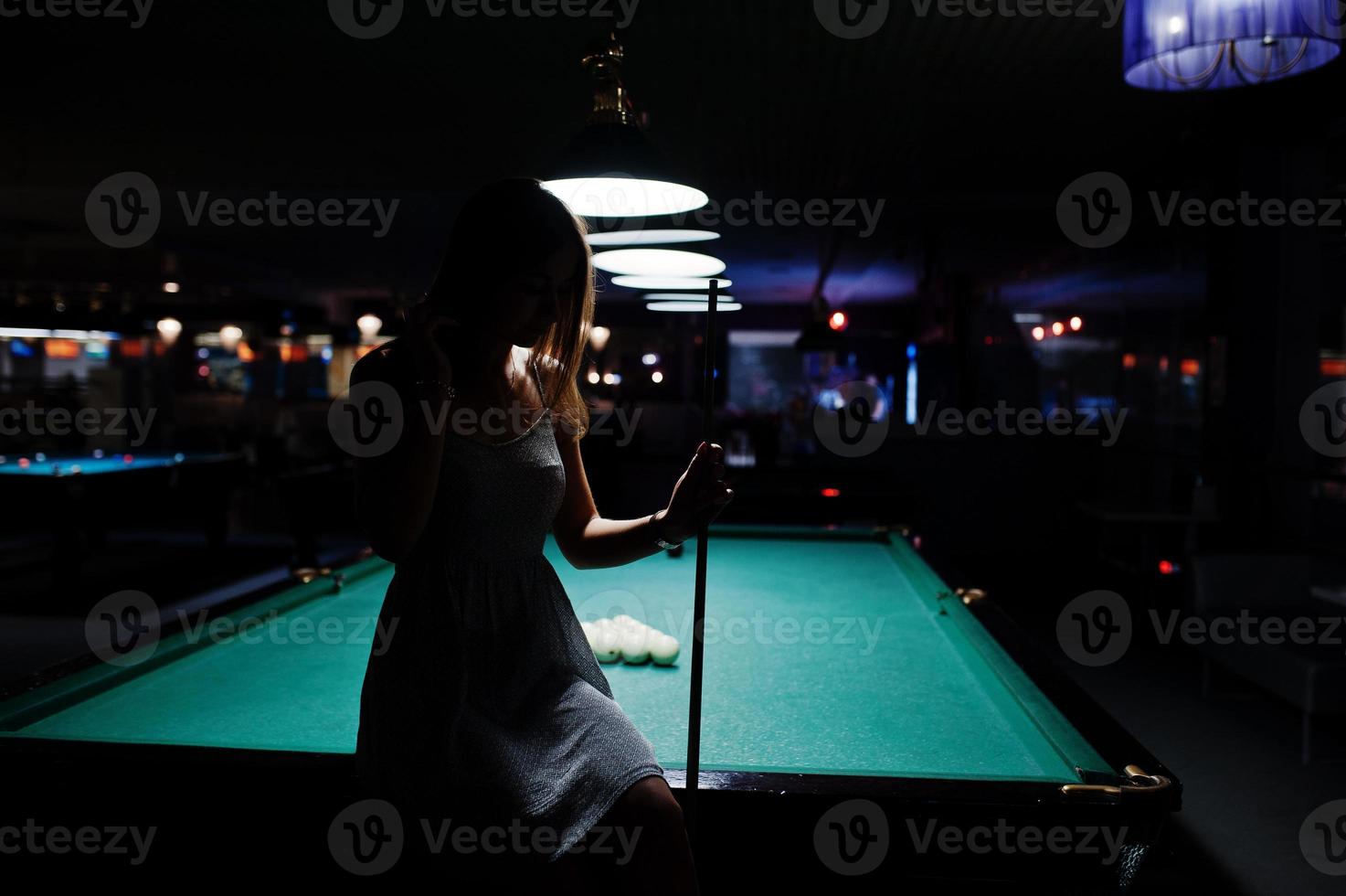 Portrait of an attractive young woman in dress sitting on the pool table and holding a cue. photo