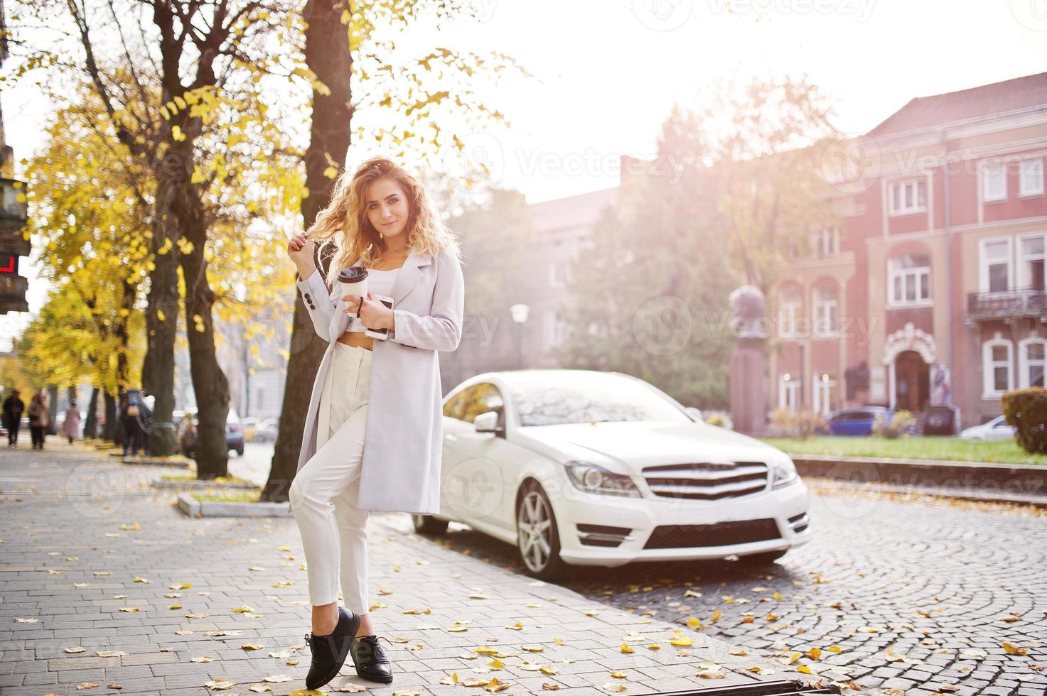 elegante modelo rubia rizada vestida de blanco con una taza de café a mano posando en la calle de otoño de la ciudad contra un auto blanco. foto