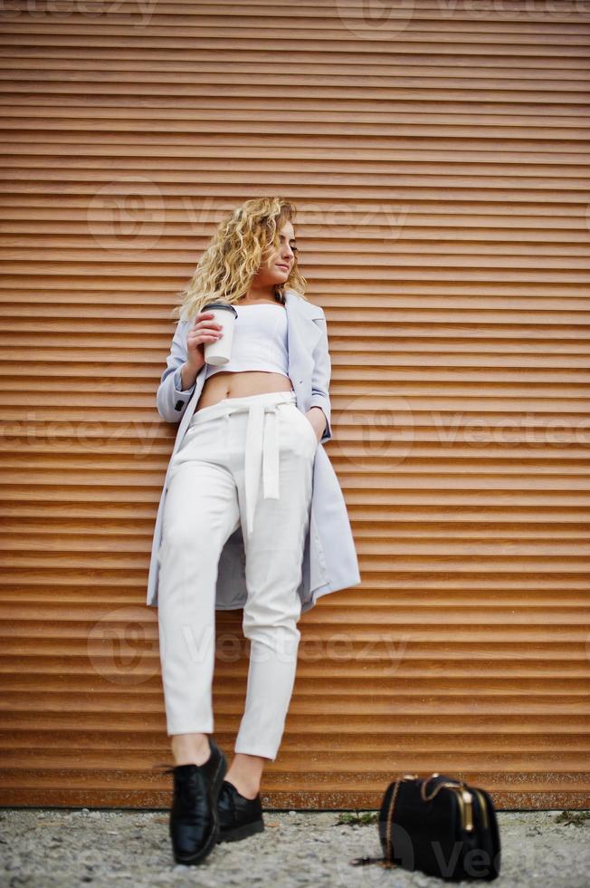 elegante modelo rubia rizada vestida de blanco con una taza de café posando contra el fondo del obturador. foto