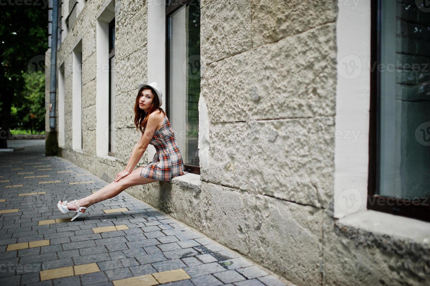 Increíbles piernas largas con tacones altos, una chica con sombrero sentada en el alféizar de la ventana en las calles de la ciudad. foto