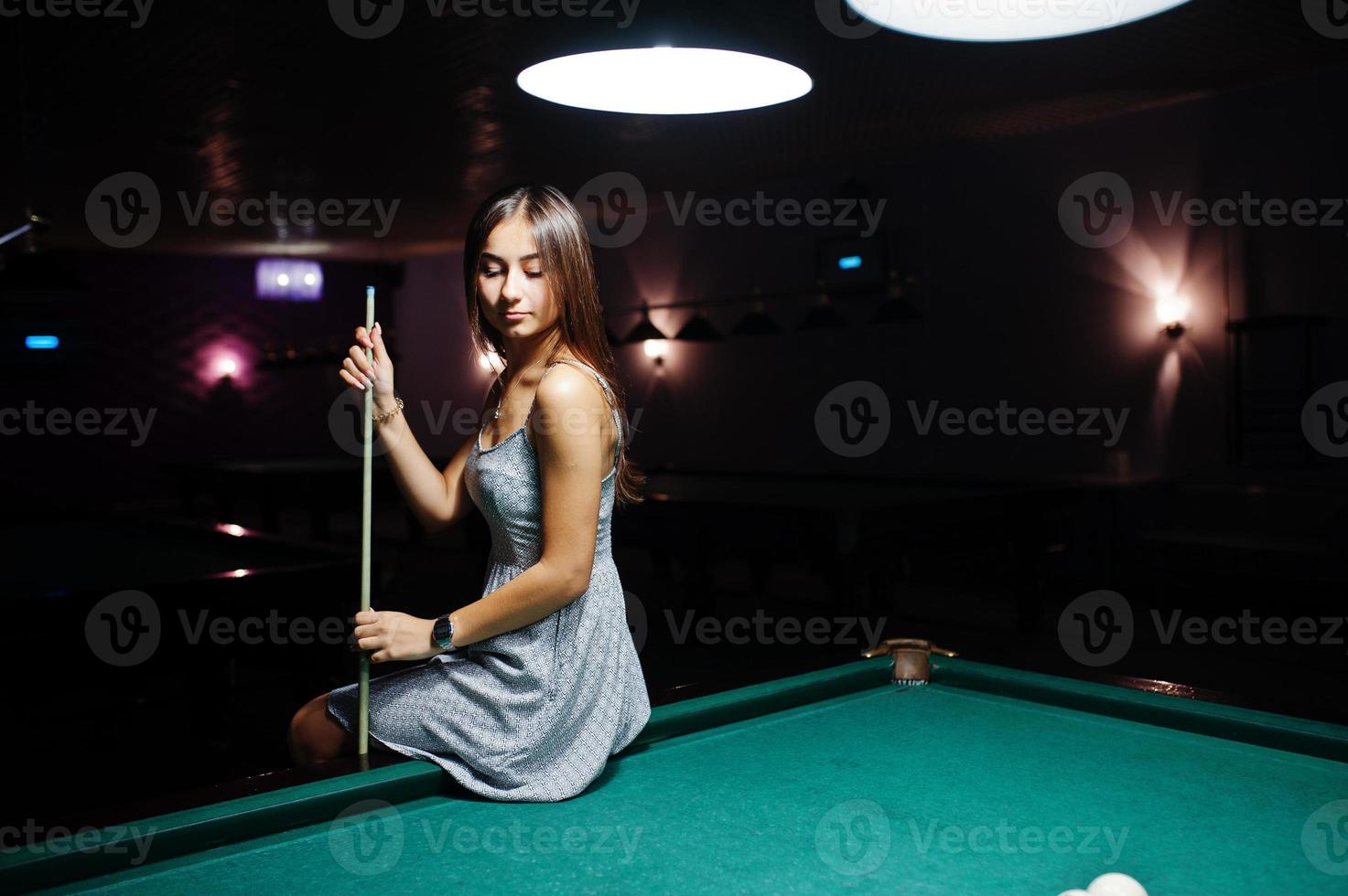 Portrait of an attractive young woman in dress sitting on the pool table and holding a cue. photo