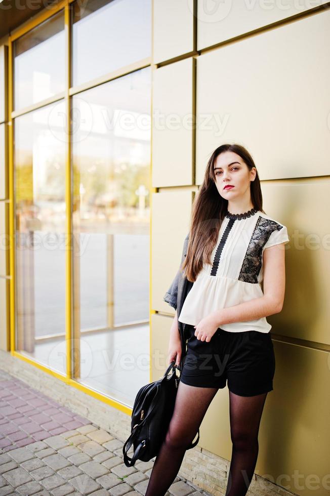 Stylish brunette girl wear on leather jacket and shorts with backpack against golden wall. photo