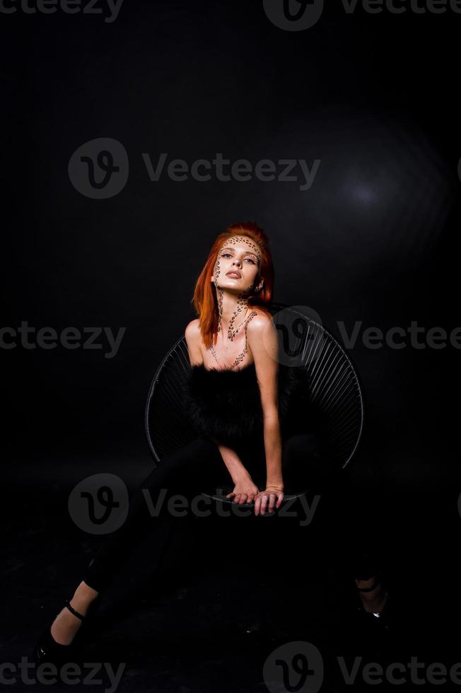 Fashion model red haired girl with originally make up like leopard predator isolated on black. Studio portrait on chair. photo