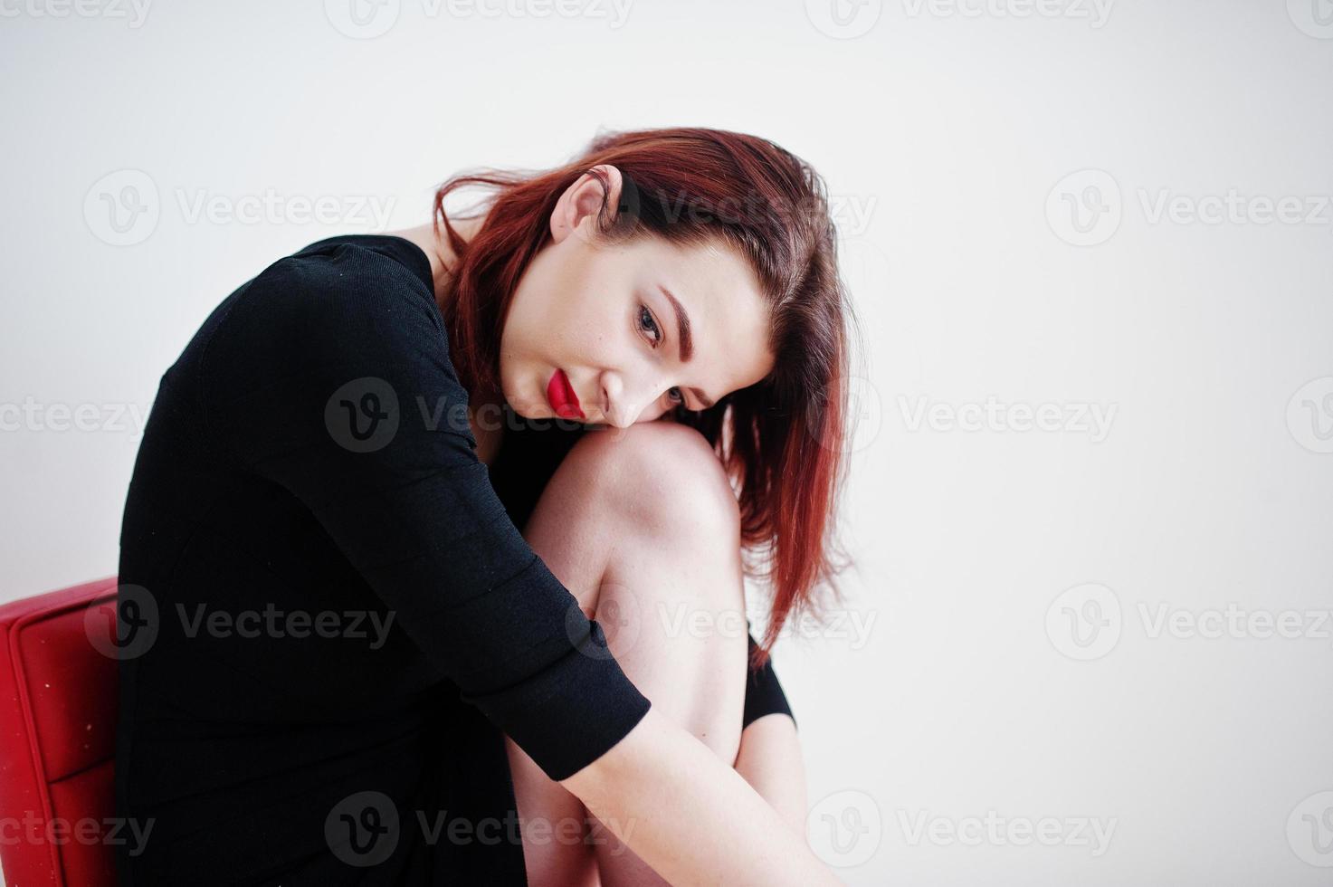 chica pelirroja con túnica negra sentada en una silla roja contra una pared blanca en una habitación vacía. foto