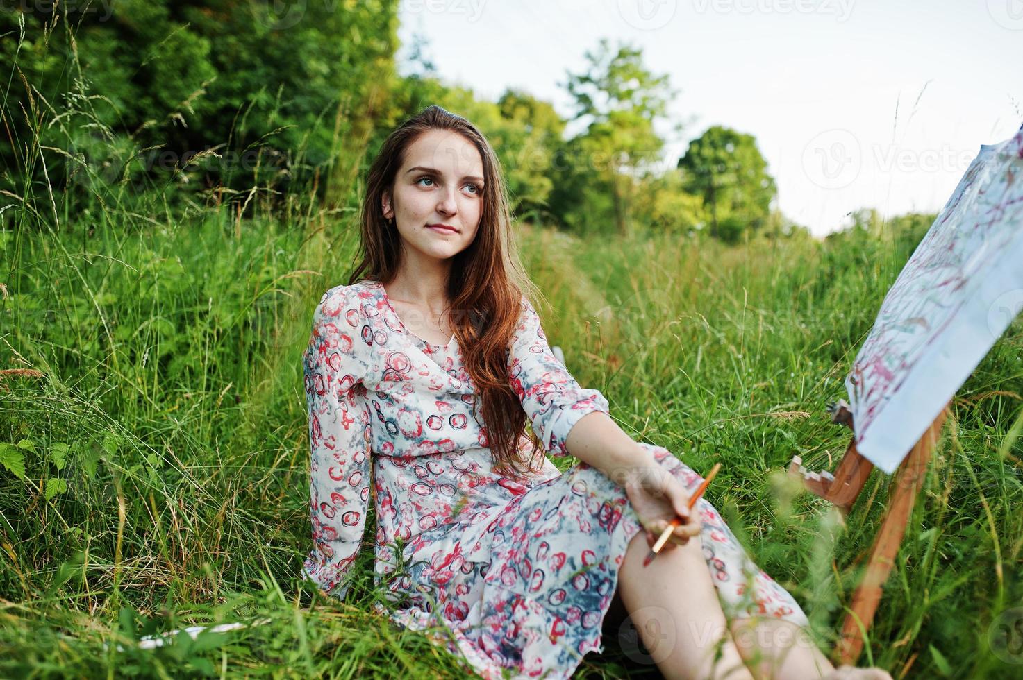 Portrait of a gorgeous happy young woman in beautiful dress sitting on the grass and painting on paper with watercolors. photo