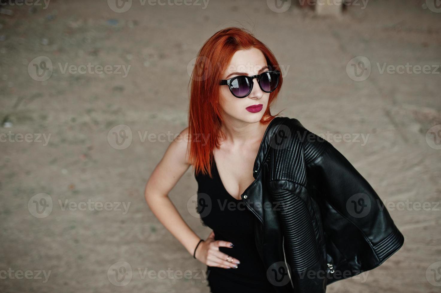 Red haired stylish girl in sunglasses wear in black, against abadoned place. photo