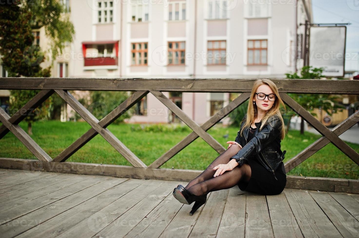Elegant blonde girl wear on black leather jacket posing at streets of town sitting on wooden. photo