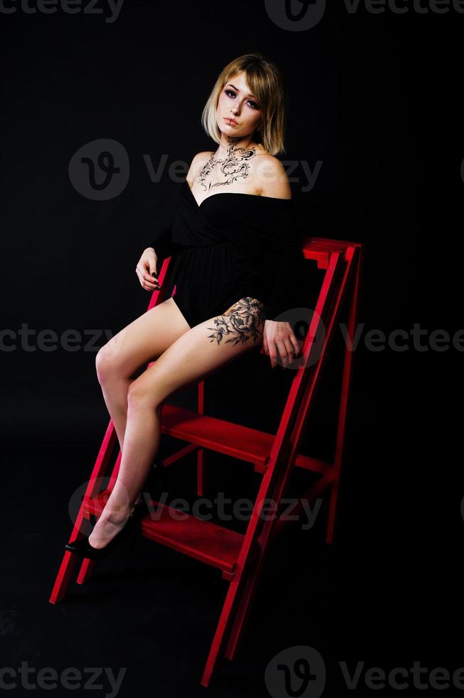 Studio portrait of blonde girl with originally make up on neck and tattoo on thigh, wear on black dress at dark background, sitting on red ladder. photo