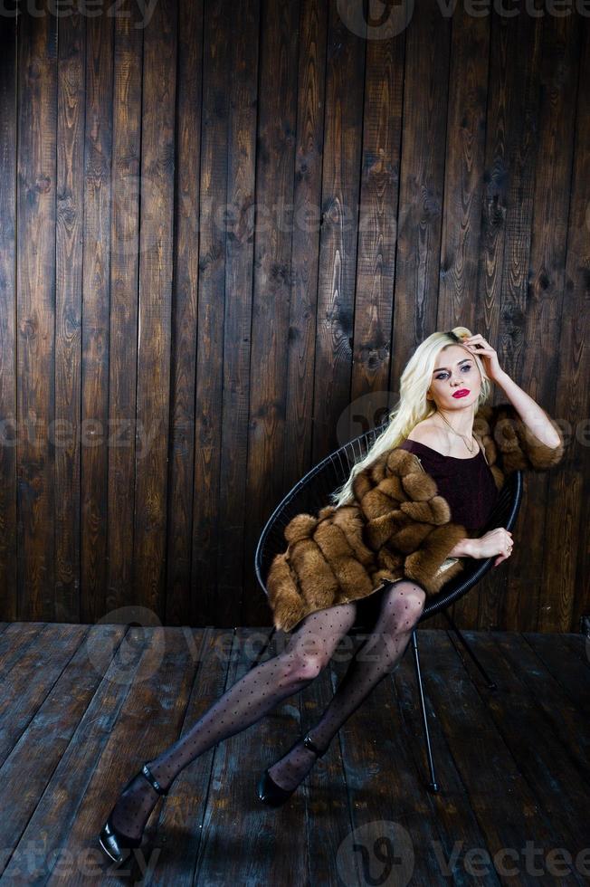 Elegante chica rubia vestida con abrigo de piel sentada en una silla en el estudio con fondo de madera. foto