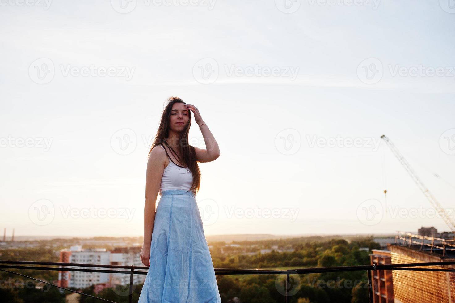 Portrait of an outstanding woman in white t-shirt and blue skirt posing on roof at sunset. photo