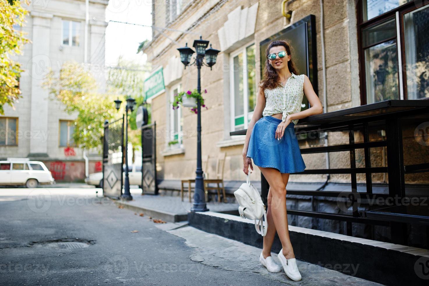 Curly stylish girl wear on blue jeans skirt, blouse and sunglasses. Portrait on streets of city. photo