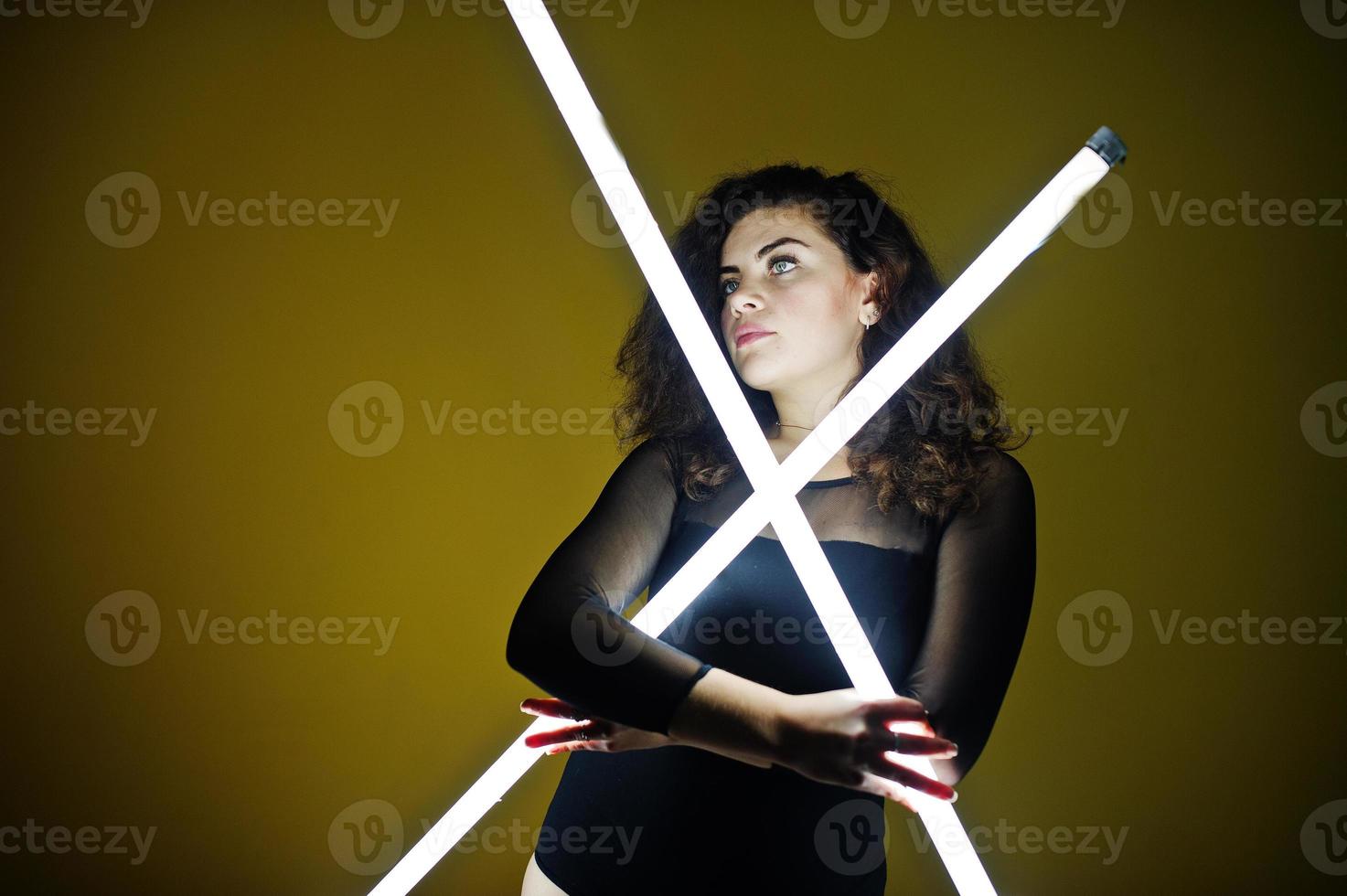 Curly brunette plus size model with two long led lamp against yellow wall. photo