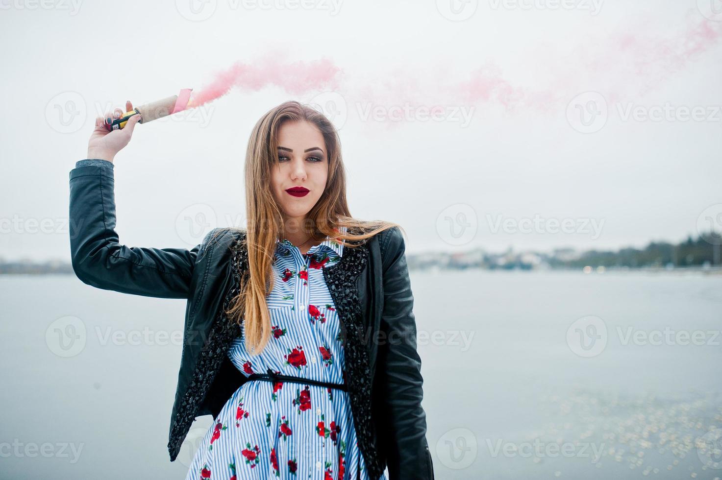 Stylish girl in leather jacket hold pink smoke flare at winter day against frozen lake. photo