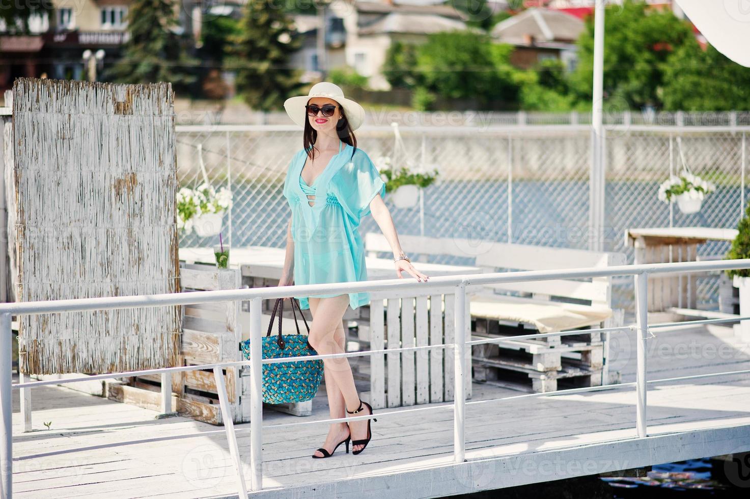 Lovely girl wearing transparent dress, hat and sunglasses walking on the quay by the lake with her bag. photo