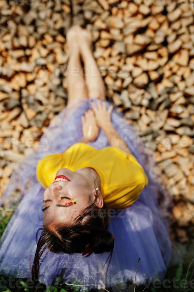 Young funny girl with bright make-up, like fairytale princess, wear on yellow shirt and violet skirt lying against wooden background. photo