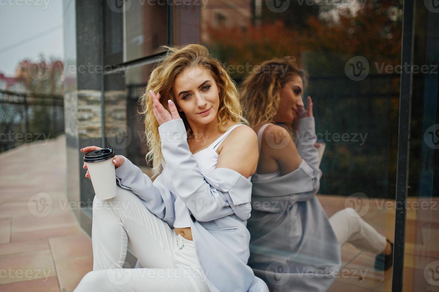 elegante modelo rubia rizada vestida de blanco con una taza de café en la mano posando contra una ventana grande. foto