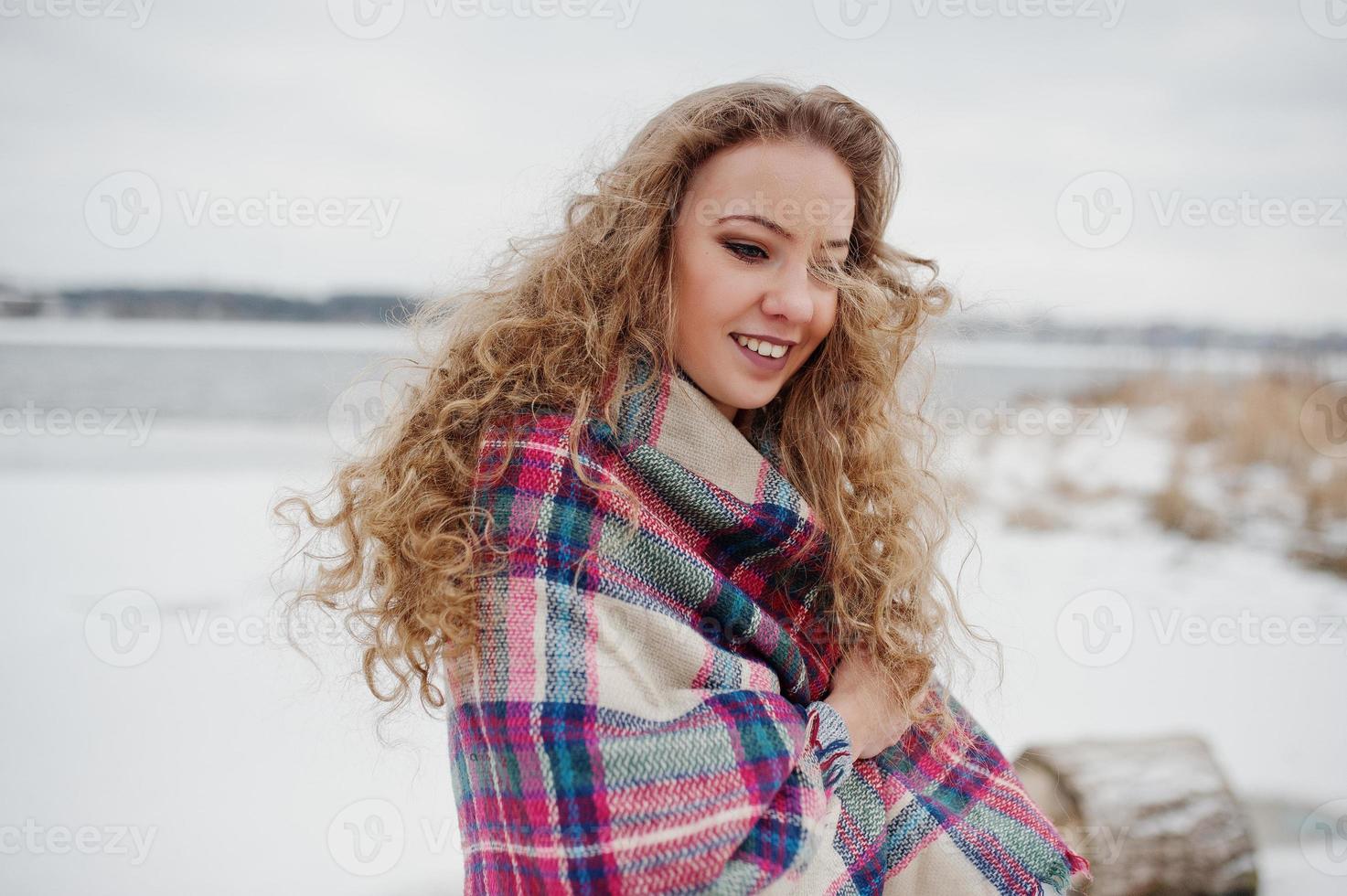 Curly blonde girl in checkered plaid against frozen lake at winter day. photo