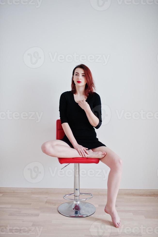 Red haired girl on black dress tunic sitting on red chair against white wall at empty room. photo