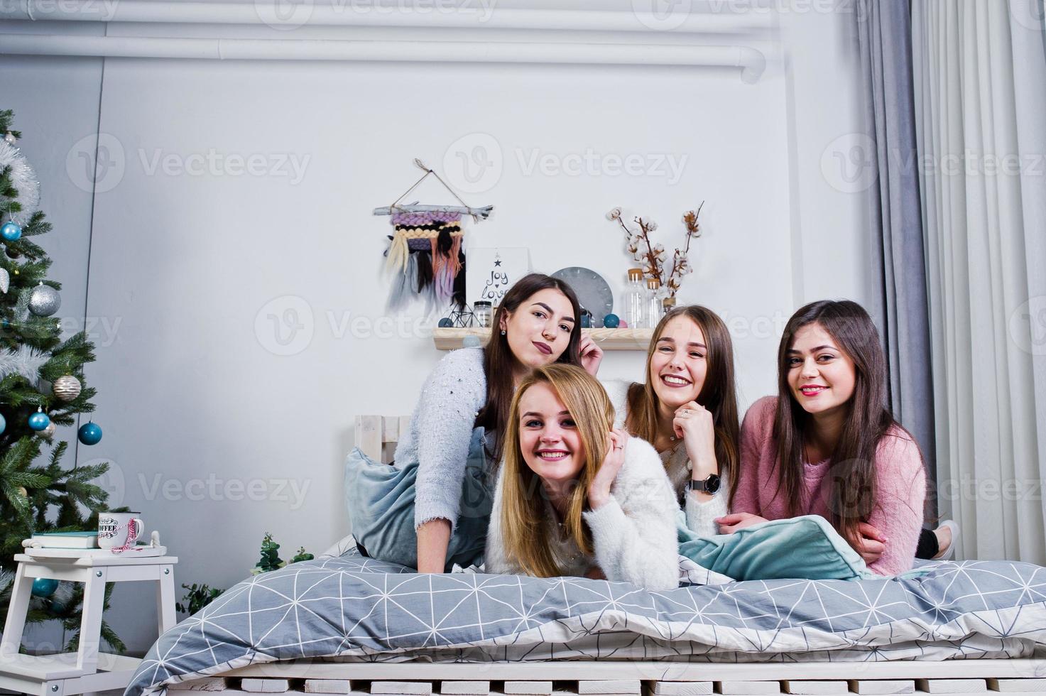 Four cute friends girls wear on warm sweaters and black pants on bed at new year decorated room on studio, play with pillows. photo