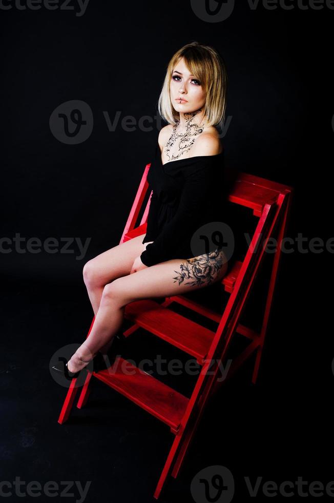 retrato de estudio de una chica rubia con maquillaje original en el cuello y tatuaje en el muslo, vestida con un vestido negro de fondo oscuro, sentada en una escalera roja. foto