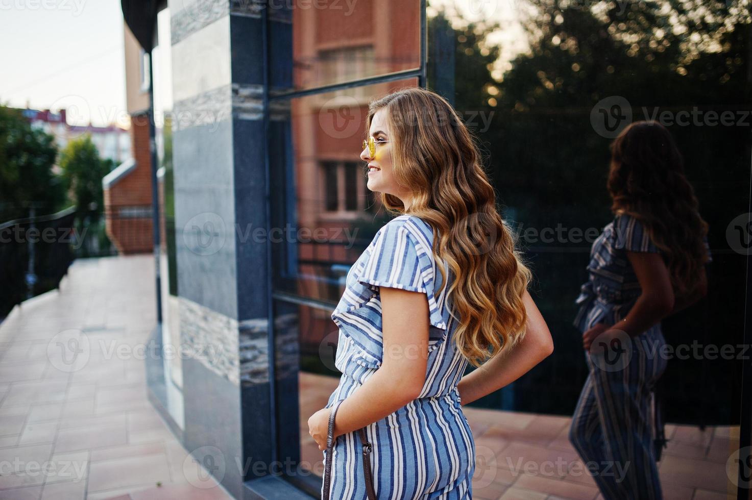 Portrait of a perfect young woman wearing striped overall and yellow sunglasses poses with her handbag on a balcony of a modern building in a town. photo