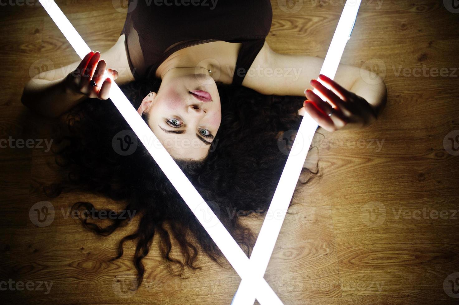 Curly brunette plus size model with two long led lamp at black room lying on flat. photo