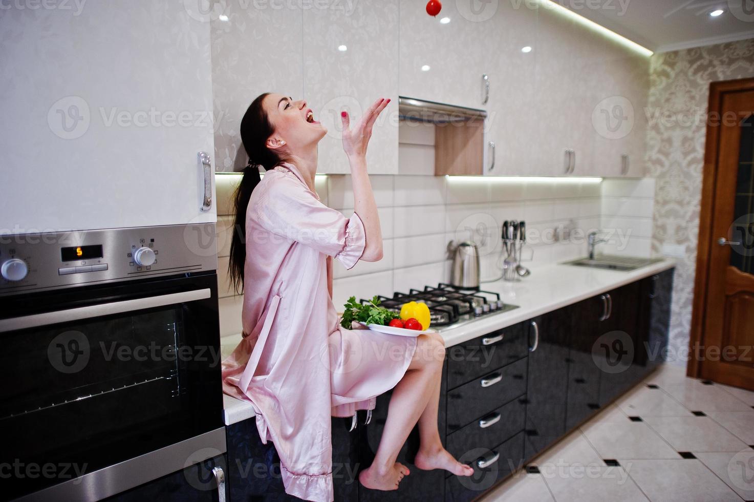 Portrait of a beautiful young woman in pink robe posing with a tomato in her kitchen. photo