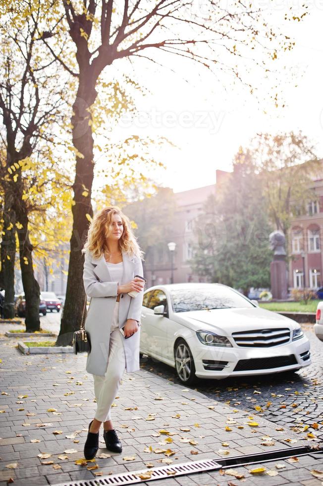 Stylish curly blonde model girl wear on white with cup of coffee at hand posing at autumn street of city against white car. photo