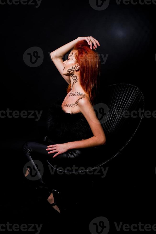 Fashion model red haired girl with originally make up like leopard predator isolated on black. Studio portrait on chair. photo