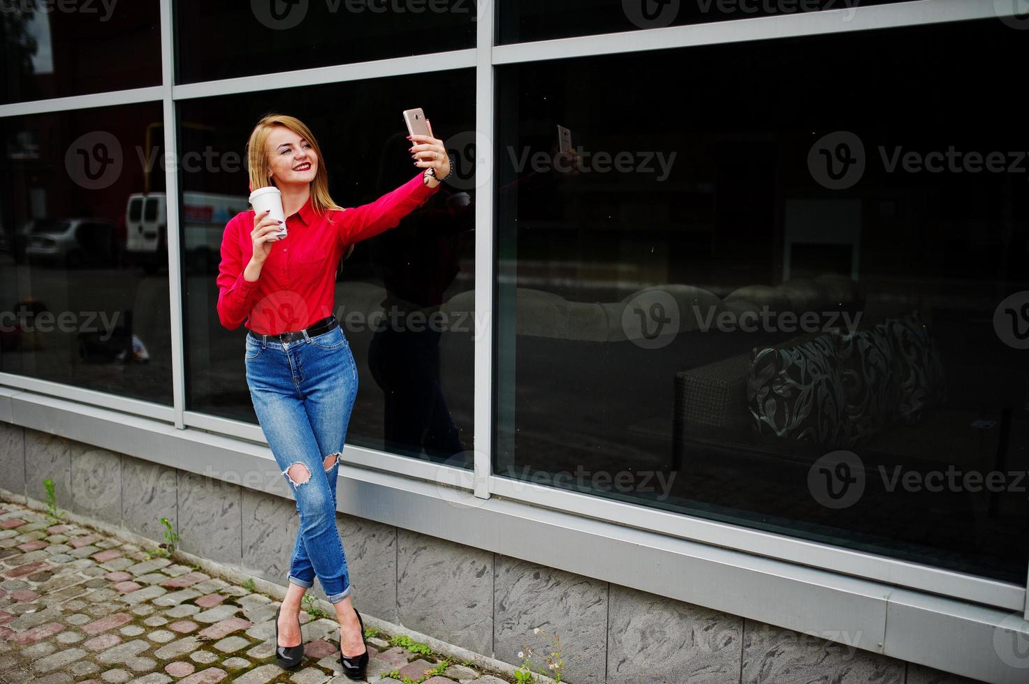 retrato de una hermosa mujer con blusa roja y jeans casuales tomando selfie en el teléfono móvil y sosteniendo una taza de café fuera del enorme centro comercial. foto