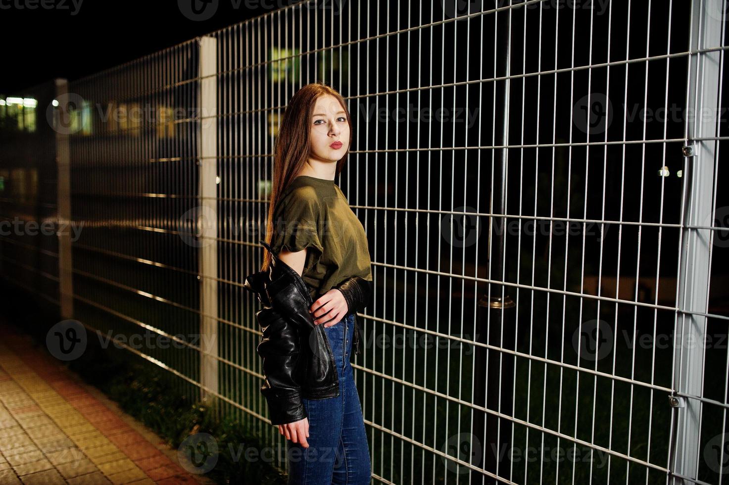 retrato nocturno de una modelo de niña vestida con jeans y chaqueta de cuero contra una verja de hierro. foto