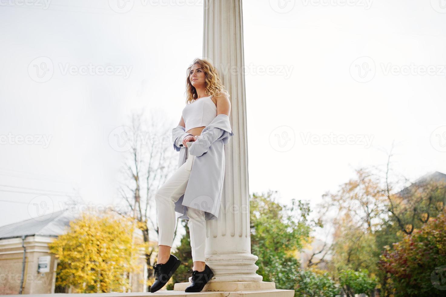 elegante modelo rubia rizada vestida de blanco posando contra la vieja casa vintage. foto