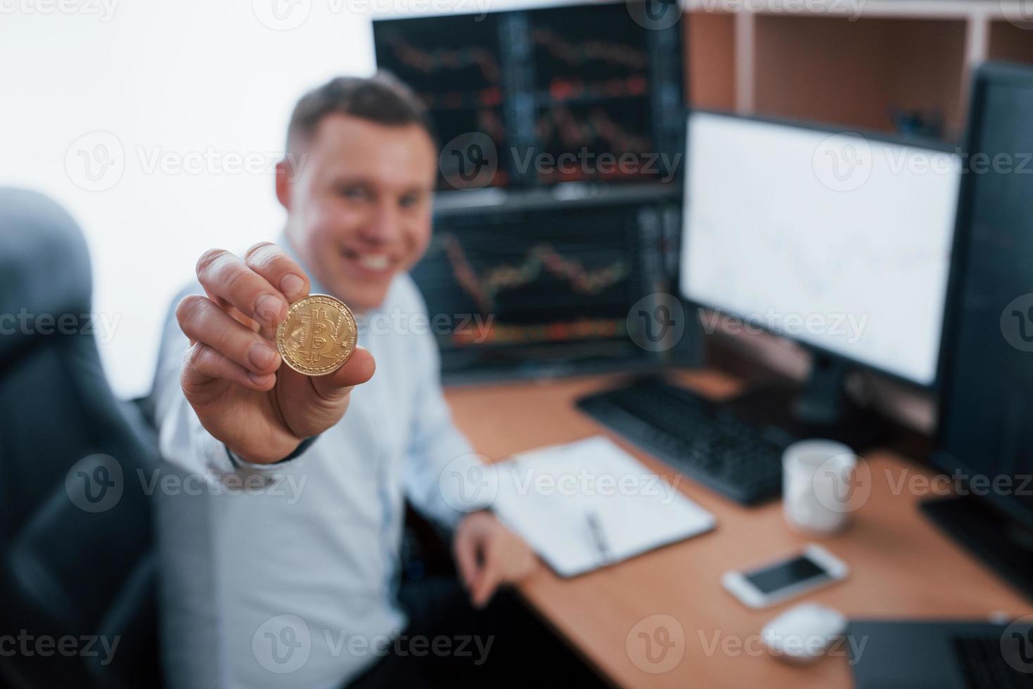 Businessman holding bitcoin in hand while sitting in modern office with many monitors with graphs photo