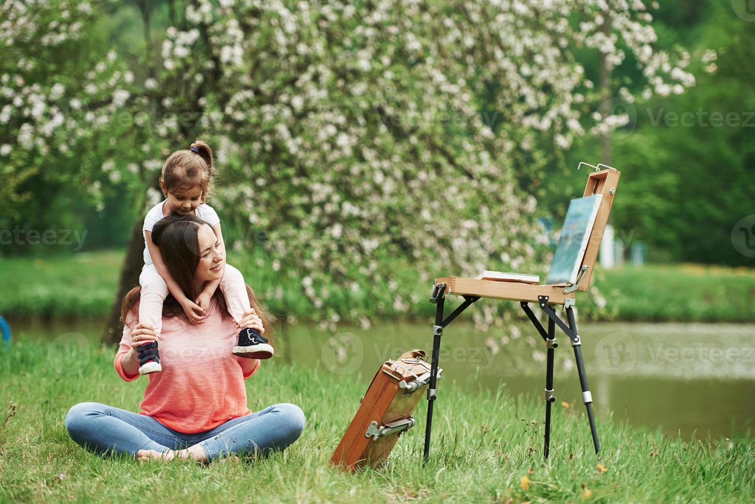 Enjoying by looking at painting. Mother and daughter sitting near easel in the park at spring time photo