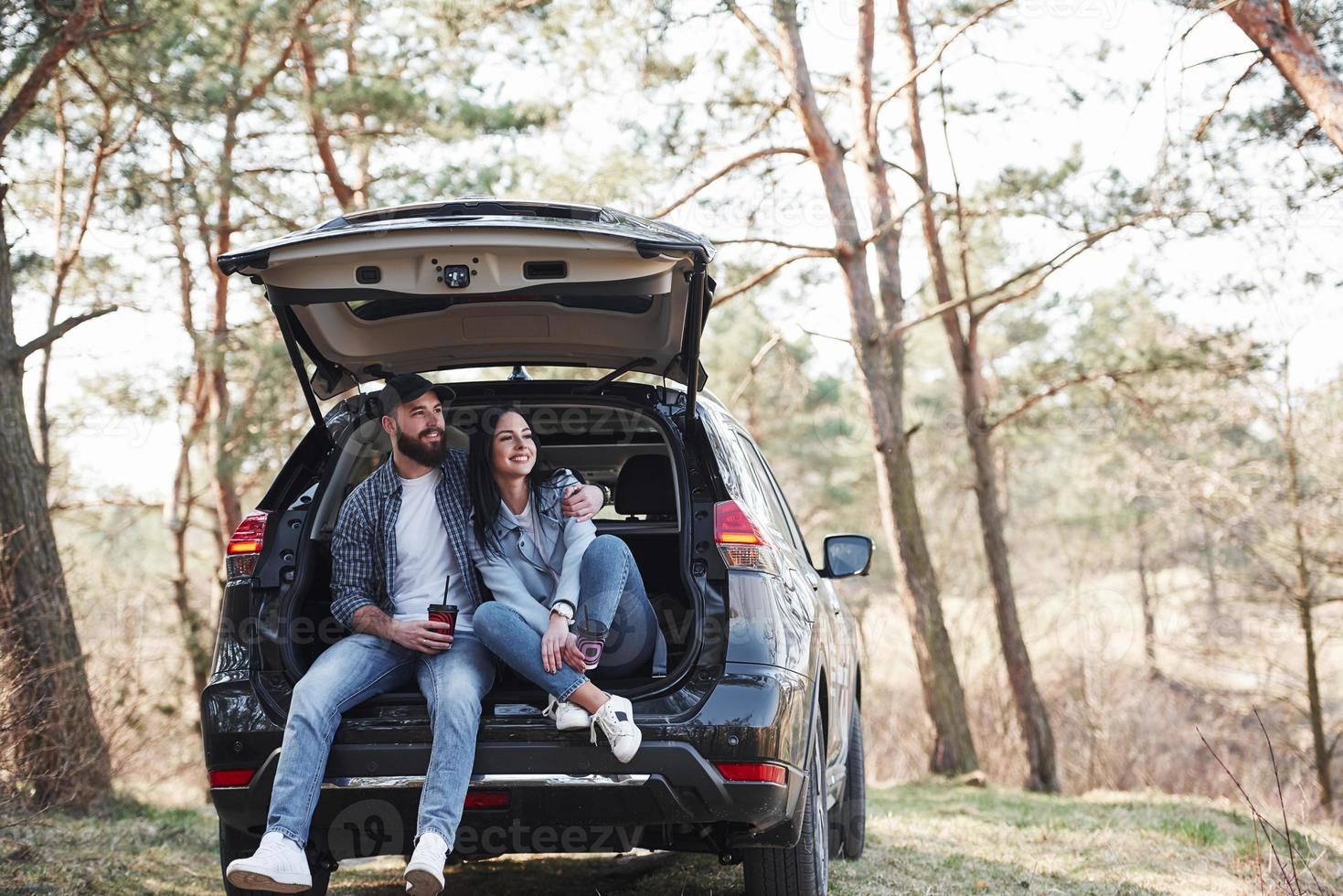 Sitting on rear part of automobile. Enjoying the nature. Couple have arrived to the forest on their brand new black car photo