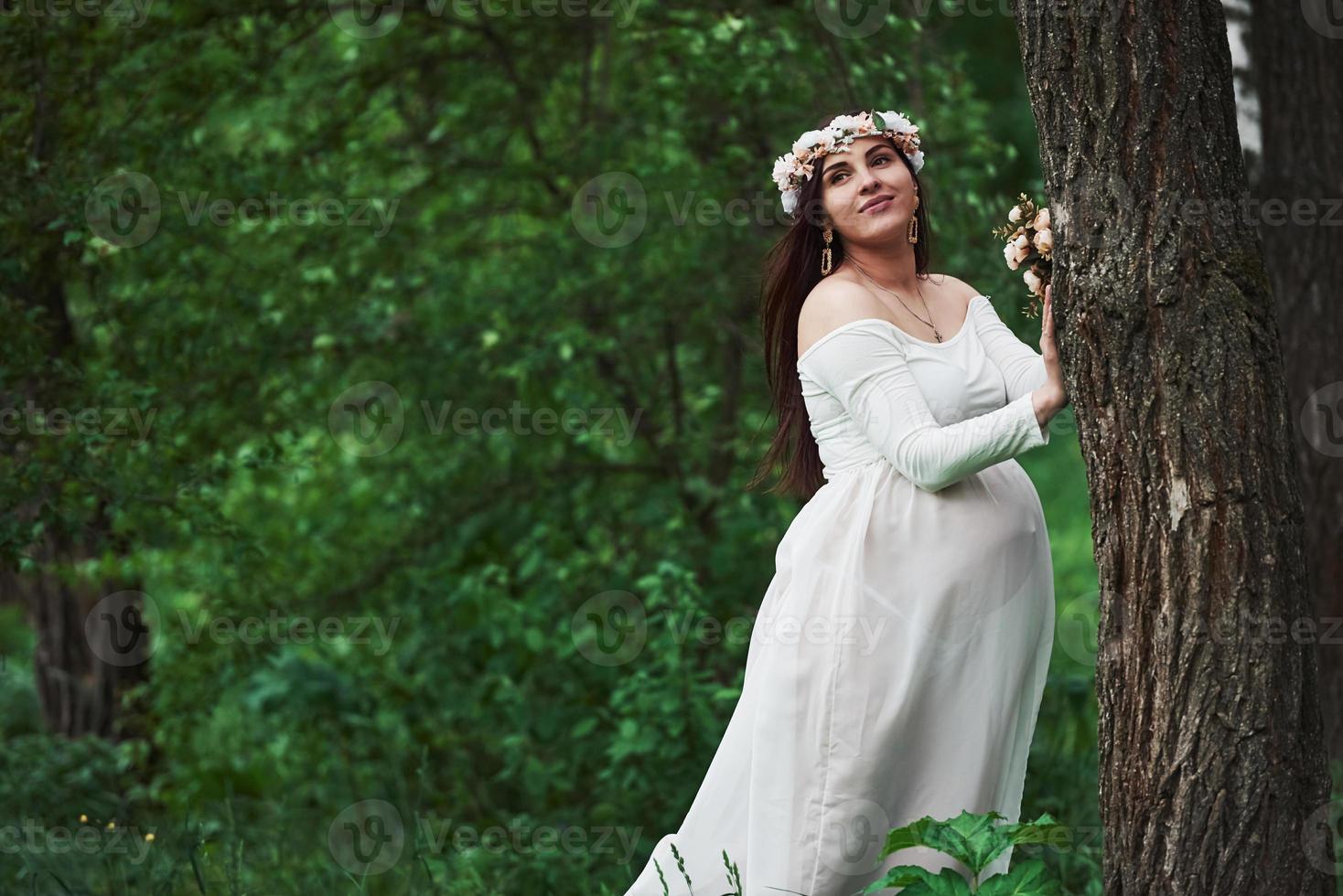 apoyado en el árbol. hermosa mujer embarazada vestida tiene un paseo al aire libre. morena positiva foto