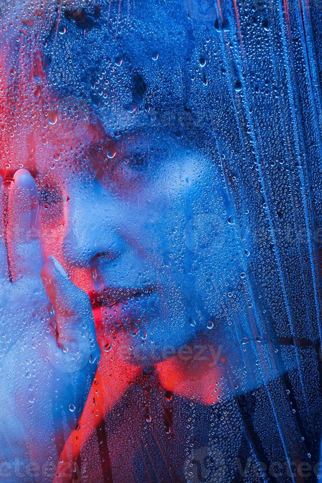 día lluvioso. foto de estudio en estudio oscuro con luz de neón. retrato de una chica hermosa detrás de un vidrio mojado