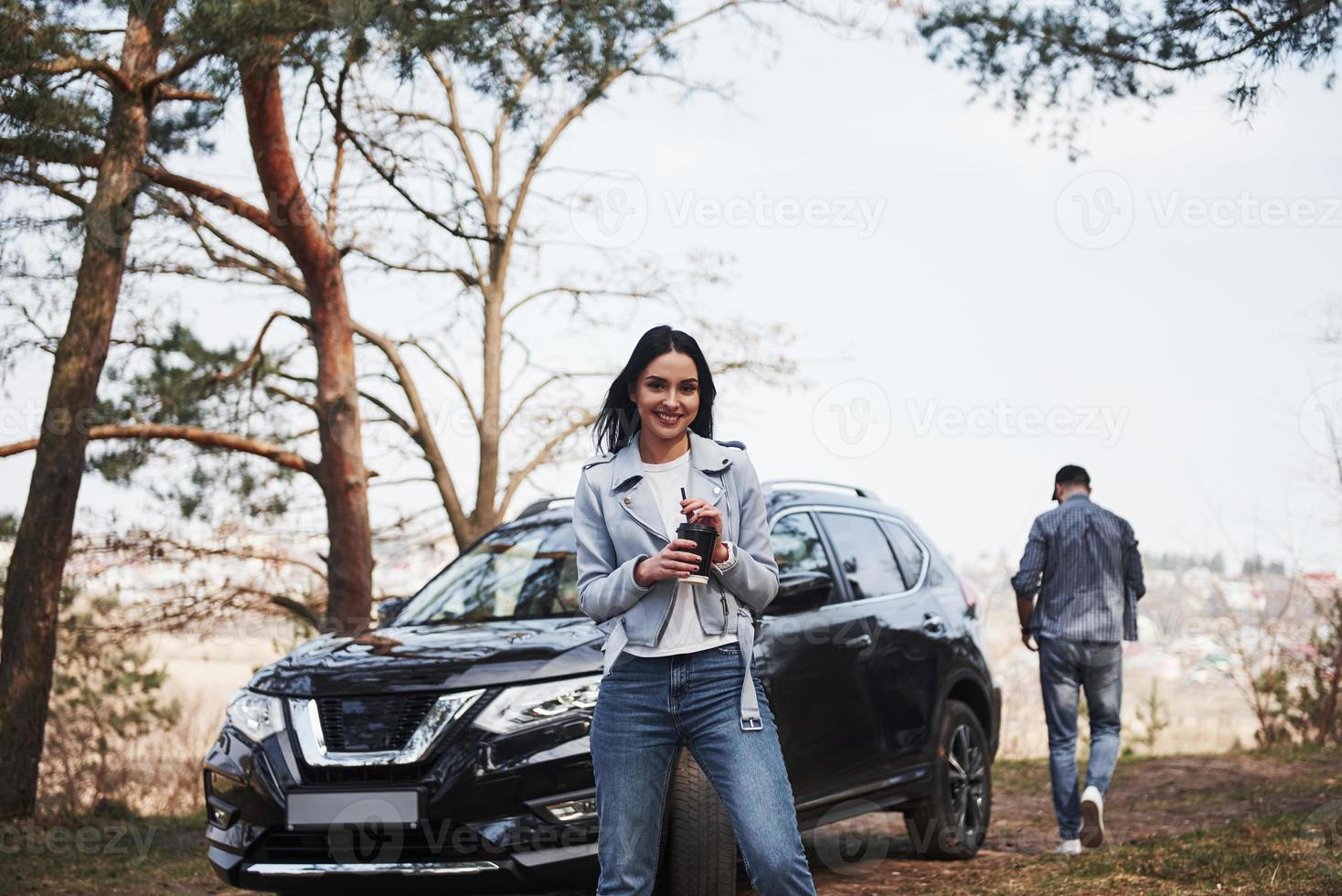 Holds cup with warm drink. Couple have arrived to the forest on their brand new black car photo