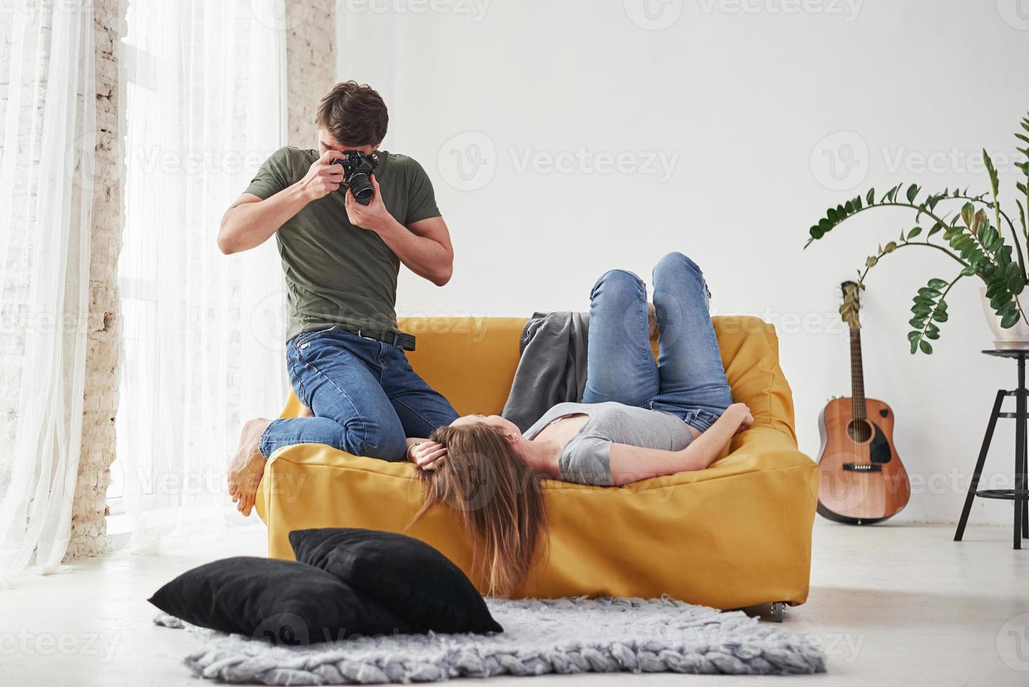 guitarra, planta y almohadas. fotógrafo tomando una foto de una joven que yacía en el sofá blanco