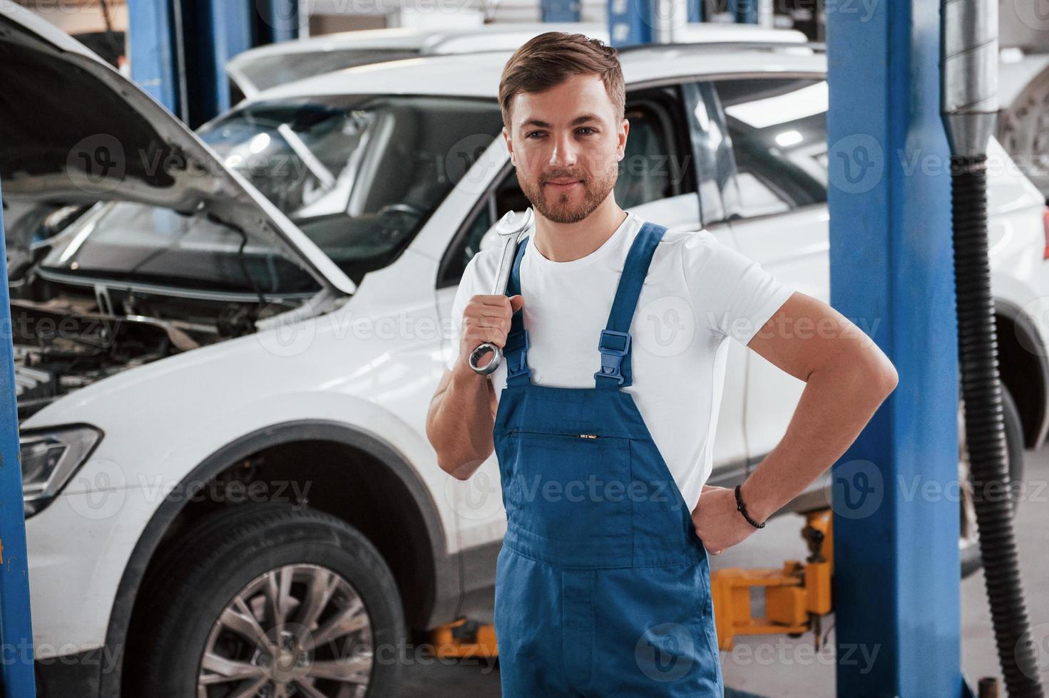 de pie con una llave en la mano. empleado en el uniforme de color azul trabaja en el salón del automóvil foto