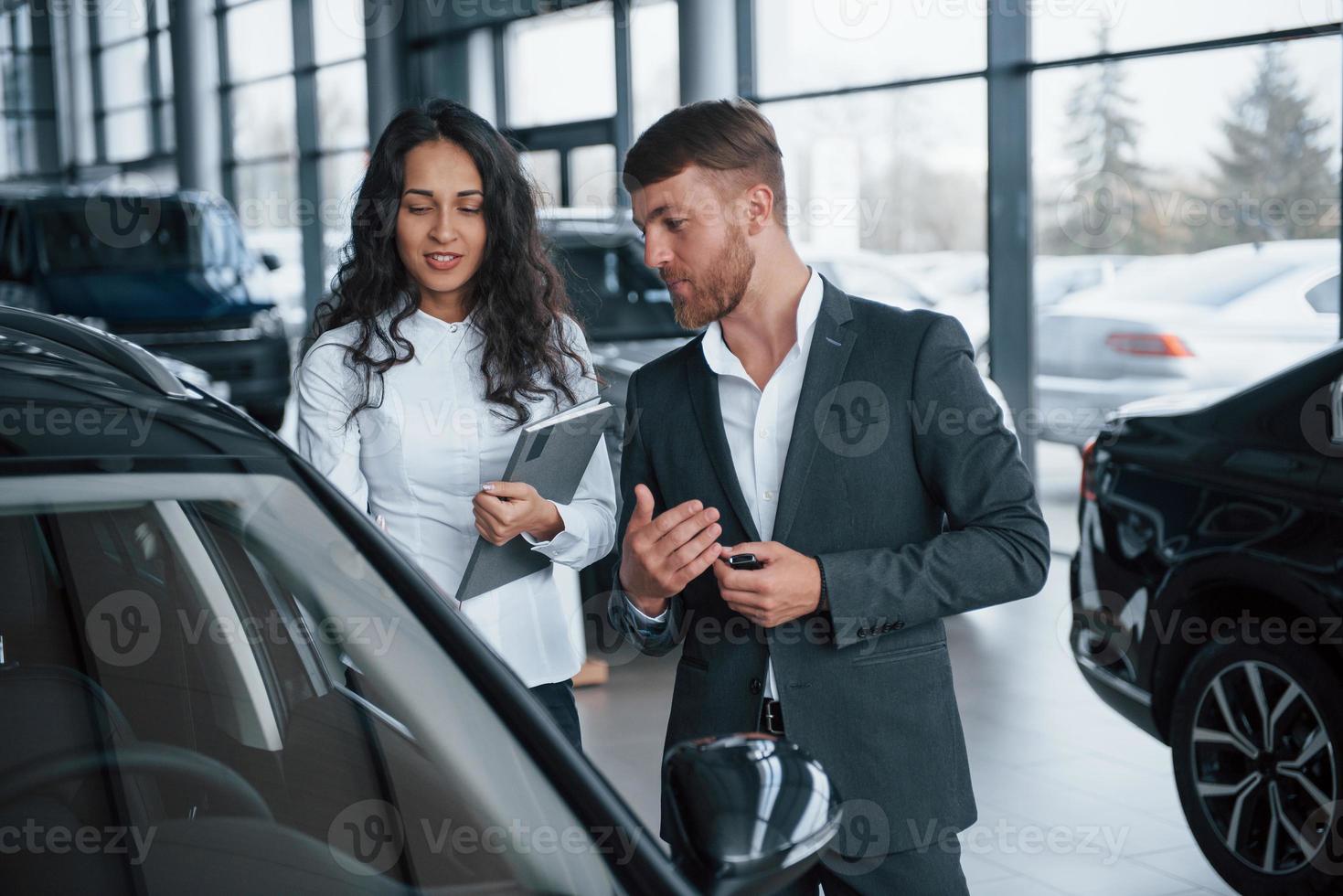 Estoy muy impresionado con ese nuevo vehículo. Cliente femenino y moderno y elegante hombre de negocios barbudo en el salón del automóvil foto