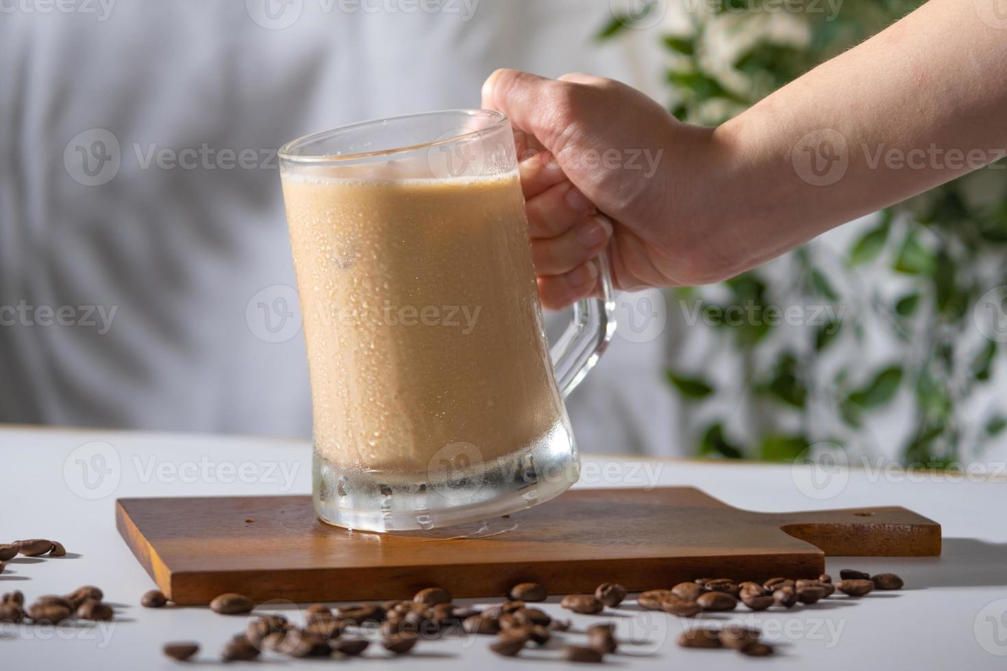 vaso de bebida de café frío con hielo y leche crema en la vieja mesa blanca en silueta abstracta sombra fondo blanco de rama de árbol de hojas naturales cayendo en la pared. Refresco de café con leche y capuchino. foto