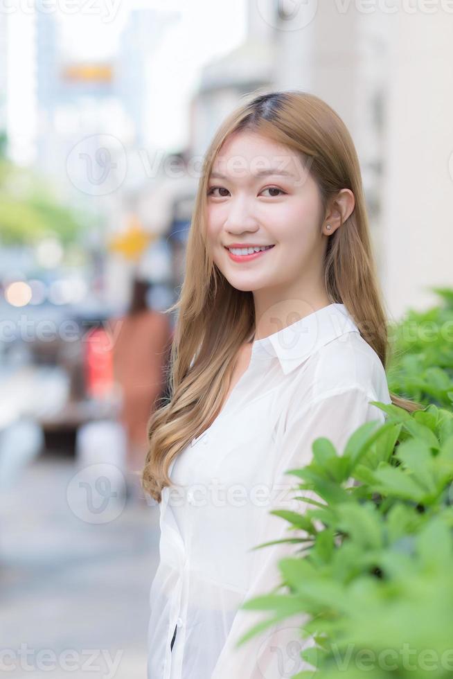 Asian beautiful woman with long bronze hair wears white long sleeve shirt and smiles happily in an urban outdoor park while looking at the camera. photo