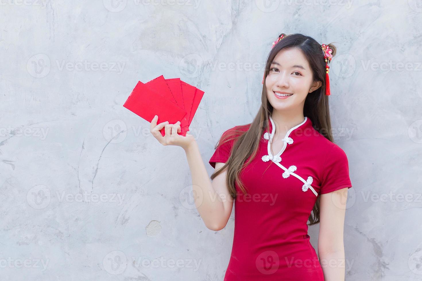 Asian beautiful girl in red dress stands and holds a red envelope with an excited expression on the grey background in Chinese new year theme. photo